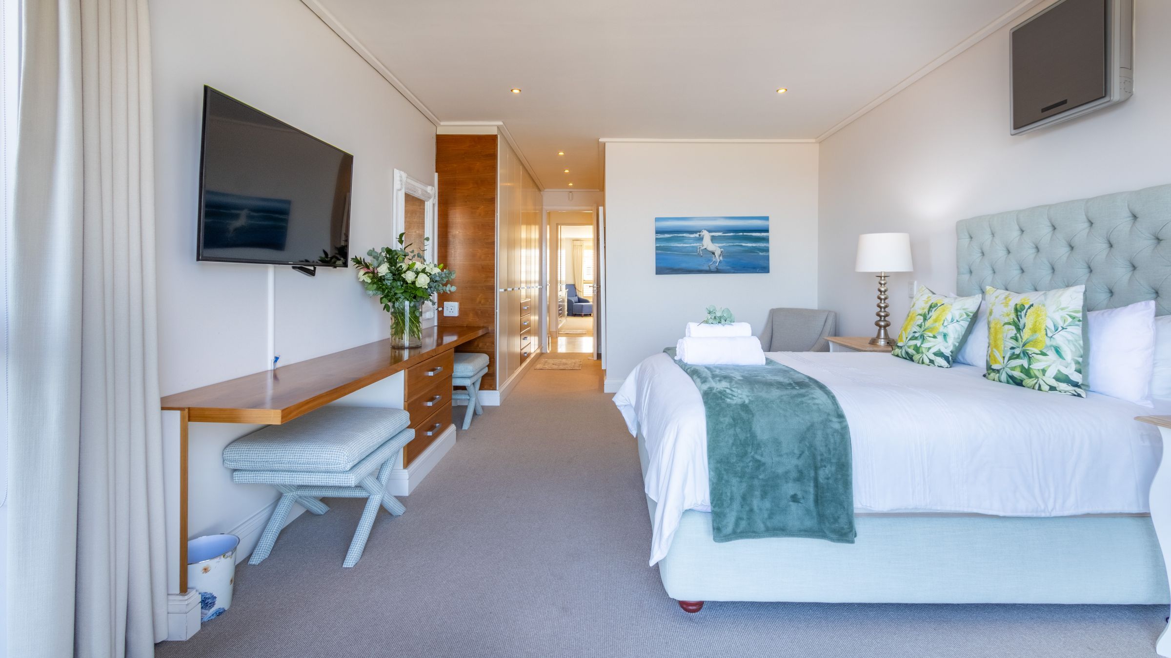 A bright hotel room featuring a large bed with floral pillows and a green throw. A wall-mounted TV, a long wooden desk with two stools, and a vase of flowers are visible. The room has neutral tones, a painting of a horse, and a doorway leading to another space.