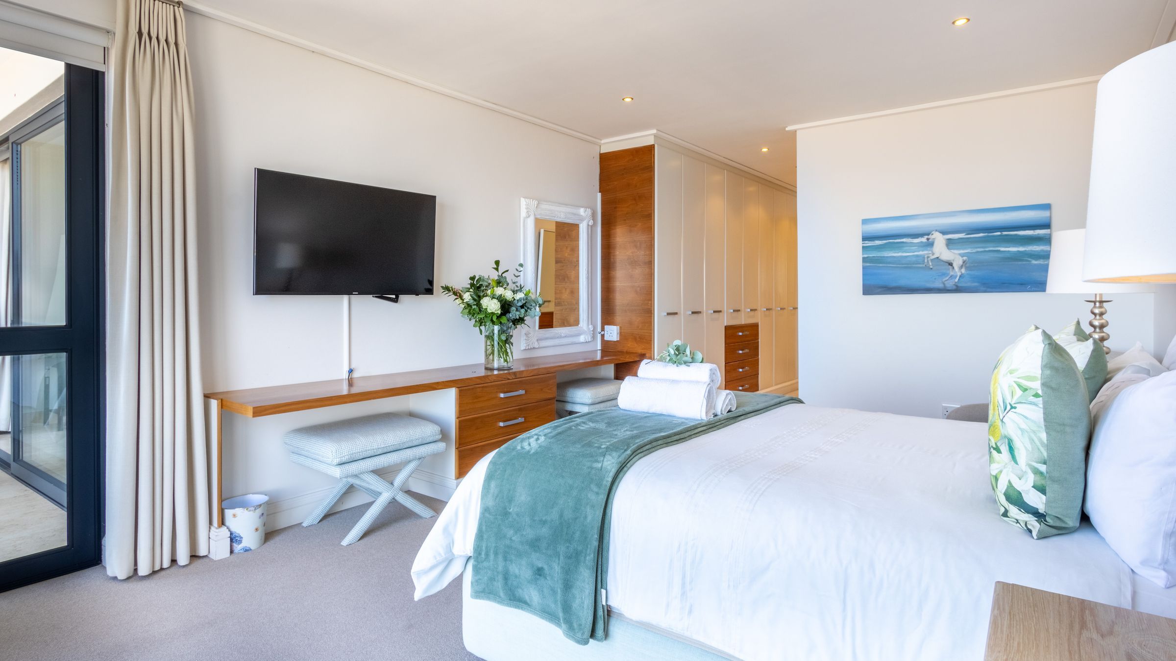 A well-lit bedroom with a large bed adorned with white and green pillows and a green throw. A mounted TV, wooden desk with a mirror, and a vase of flowers are visible. A painting of a white horse by the sea and a stack of towels add to the decor. Large windows provide natural light.