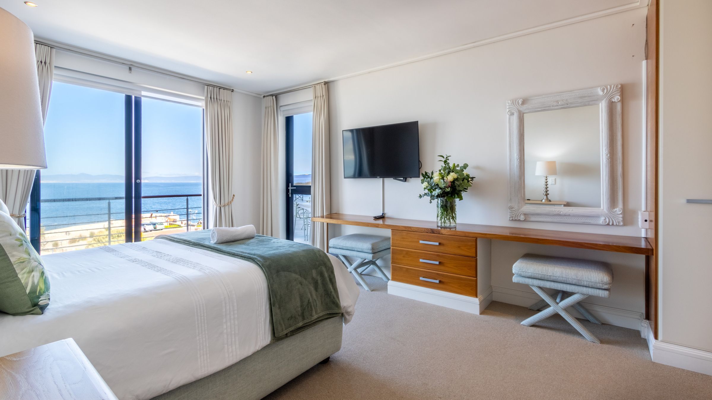 A bright bedroom featuring a large bed with white linens and green accents, facing a flat-screen TV and a wooden dresser. A mirror and a floral arrangement add elegance. Sliding glass doors open to a balcony with a sea view, framed by light curtains, enhancing the airy atmosphere.