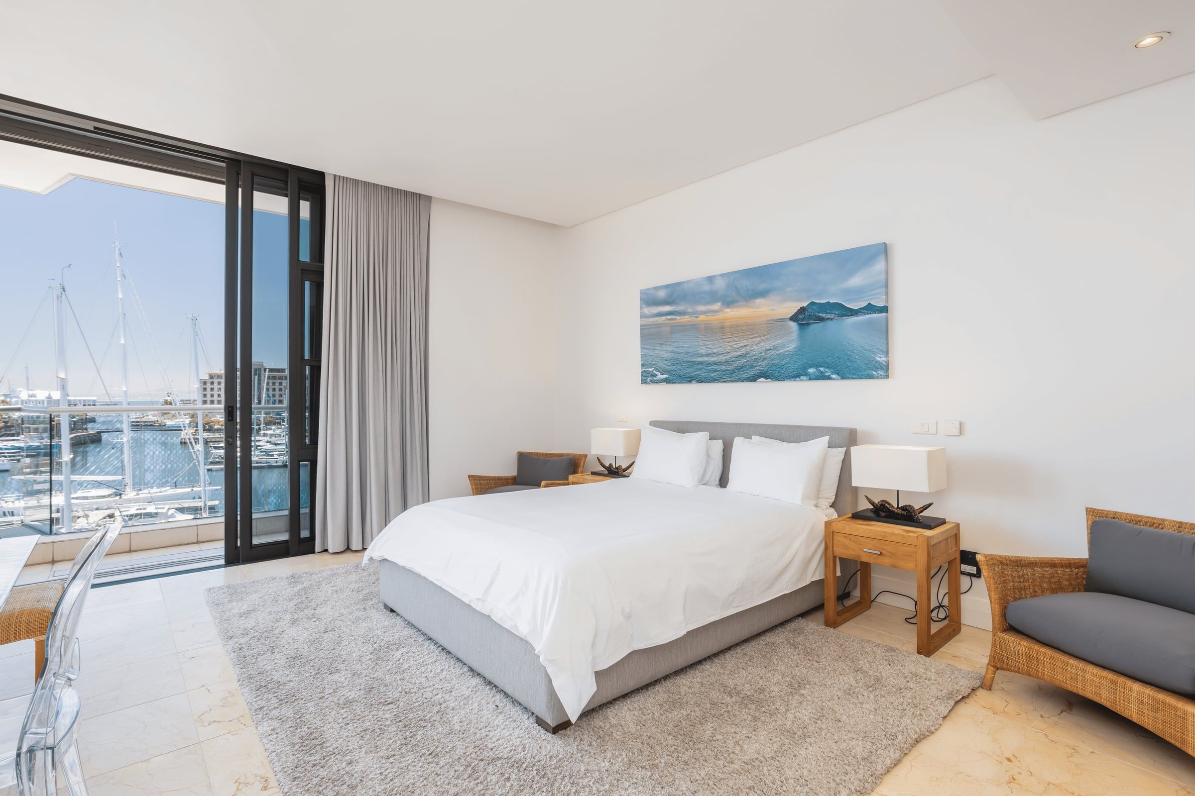 A stylish bedroom with a large bed, white linens, wooden nightstands, and lamps. A gray rug covers the floor. A glass door opens to a balcony overlooking a marina with sailboats. A landscape painting hangs above the bed.