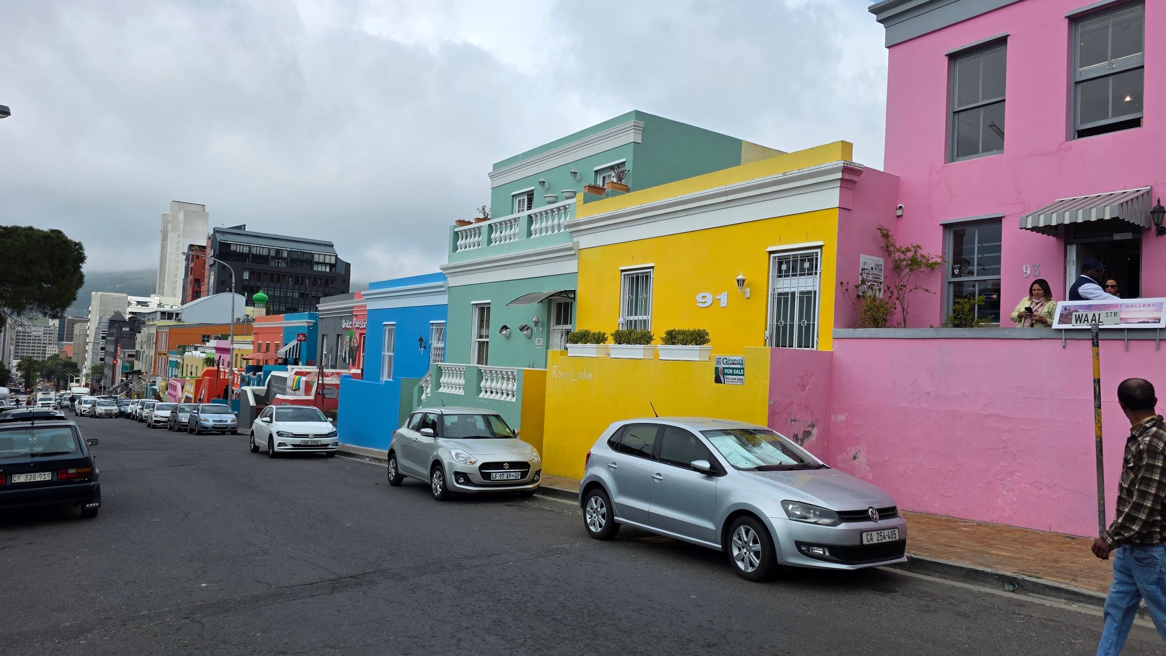 A vibrant street in Bo-Kaap, Cape Town, featuring brightly painted houses in pink, yellow, blue, and green. Cars are parked along the cobblestone road, and people engage in conversation, reflecting the rich Cape Malay heritage and community warmth.