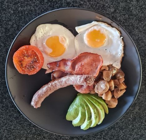 A hearty breakfast plate featuring two sunny-side-up eggs, a grilled tomato slice, crispy bacon, sautéed mushrooms, a sausage link, and sliced avocado, arranged on a dark round plate against a dark background.