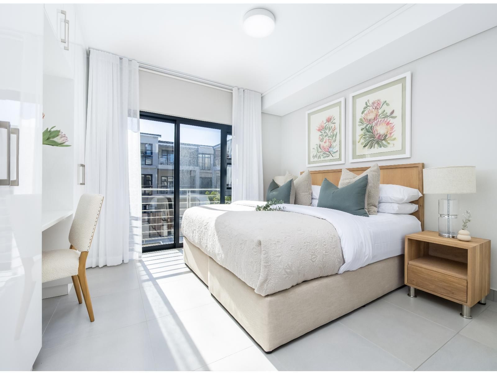 Elegant bedroom with a large bed, floral artwork, and a wooden side table. A desk with a cushioned chair is by the window leading to a balcony. Natural light enhances the room's tranquility.