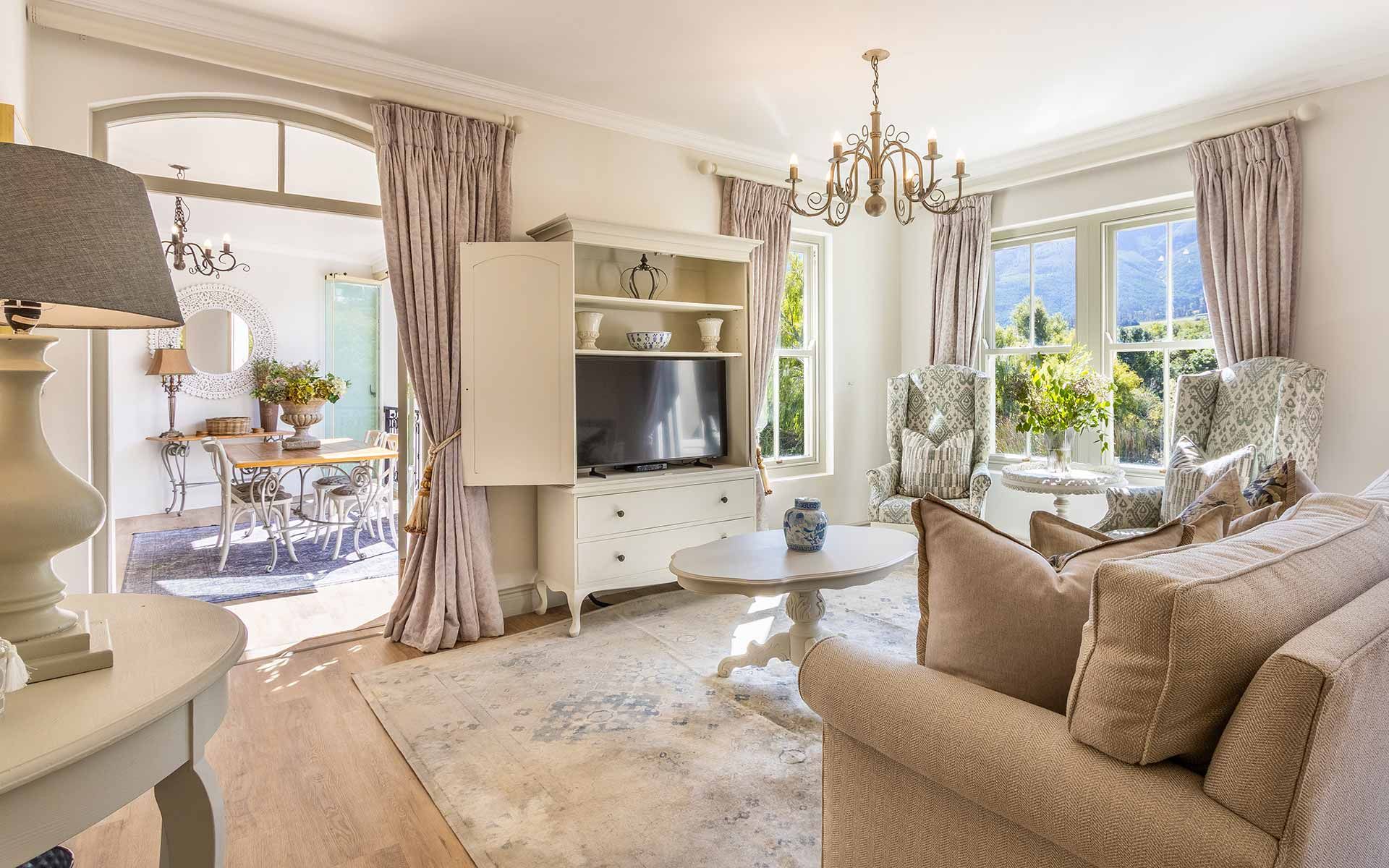 A charming sitting room with a neutral palette, featuring a beige sofa, patterned armchairs, and a round coffee table. Large windows offer views of lush greenery. A dining area with a round table and chairs is visible through an archway, complemented by elegant chandeliers and soft drapes.