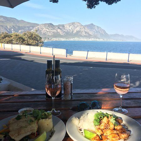 Two plated dishes with fresh ingredients sit on a wooden table overlooking the ocean and mountains. A glass of rosé wine accompanies the meal, enhancing the dining experience in a picturesque setting.