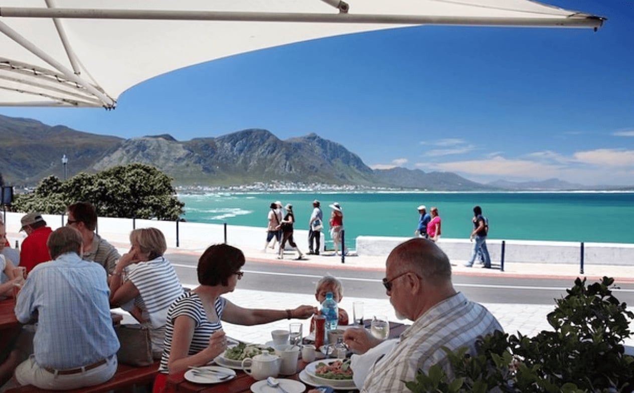 A group of people enjoy a meal under a shaded patio near the sea. In the background, people stroll along the promenade against a backdrop of turquoise water and distant mountains under a clear blue sky.