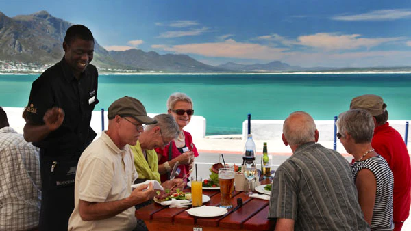 A group enjoys a meal at Burgundy Restaurant in Hermanus, South Africa. The outdoor seating offers stunning ocean views. A waiter smiles as he serves the guests, who are seated at a wooden table with various dishes and drinks. The scene is vibrant and inviting.