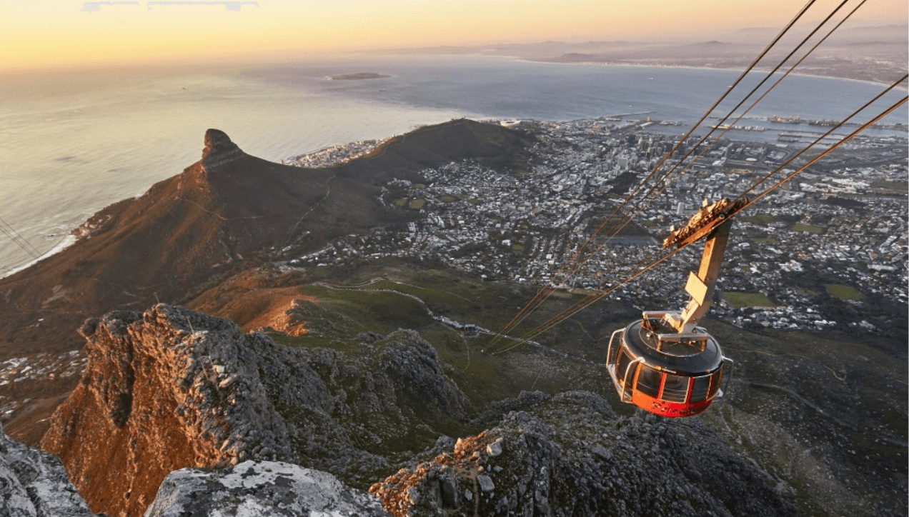 A breathtaking view from Table Mountain showcasing the aerial cableway descending towards Cape Town. The flat-topped summit is visible with the city sprawling below, bordered by the ocean. The scene captures the mountain's natural beauty and its status as a UNESCO site.