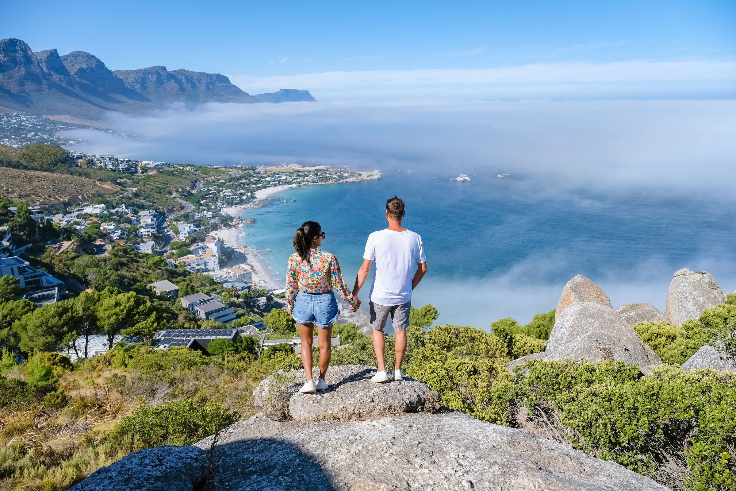 A couple stands on a rock, holding hands, overlooking a scenic coastal bay with clear blue water, surrounded by green hills and distant mountains under a bright blue sky. The landscape includes a small town with houses and roads along the shoreline.