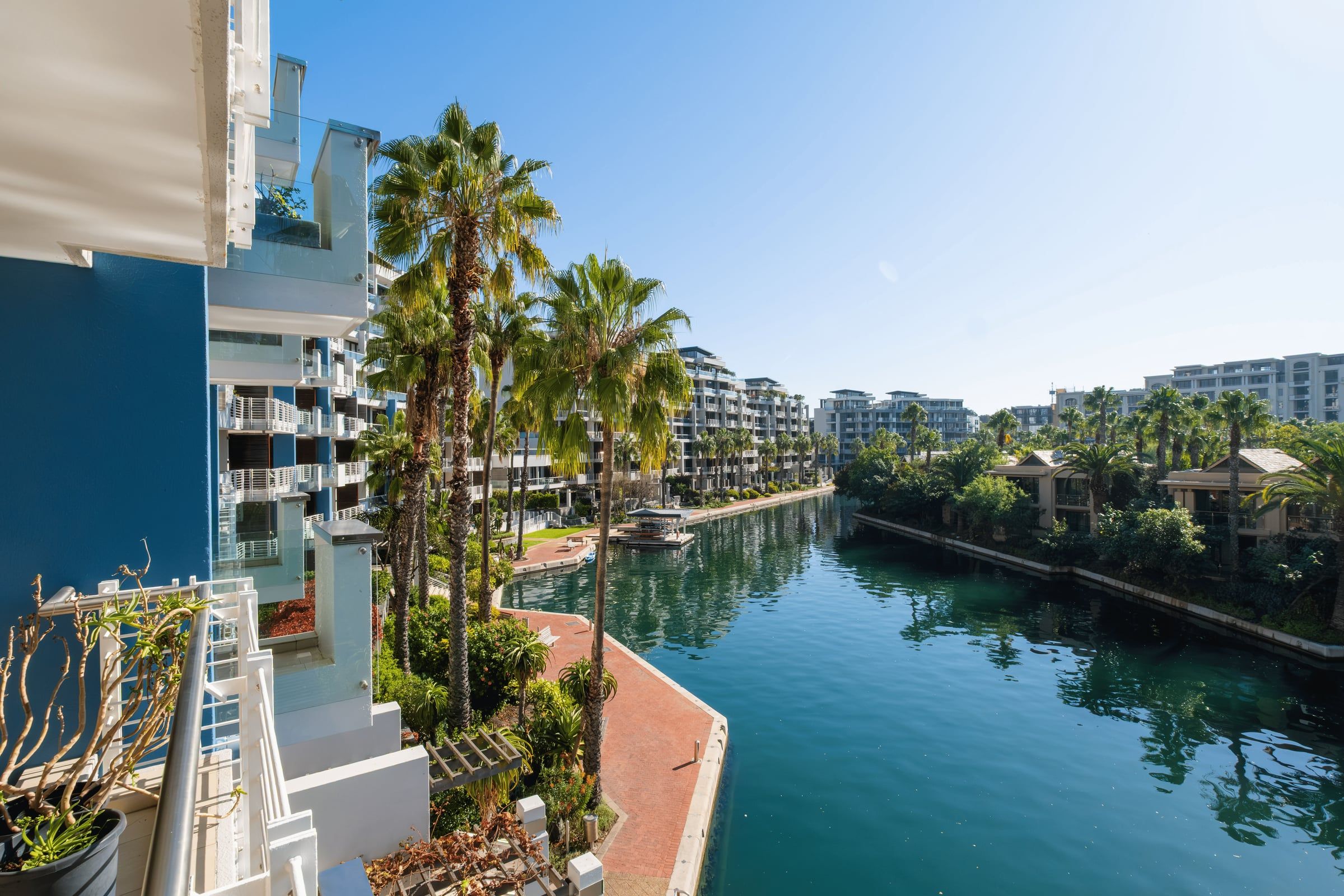 View of modern waterfront apartments with blue facades, palm trees, and a calm canal. The scene is sunny with clear skies, showcasing lush greenery and reflections in the water, creating a serene atmosphere.
