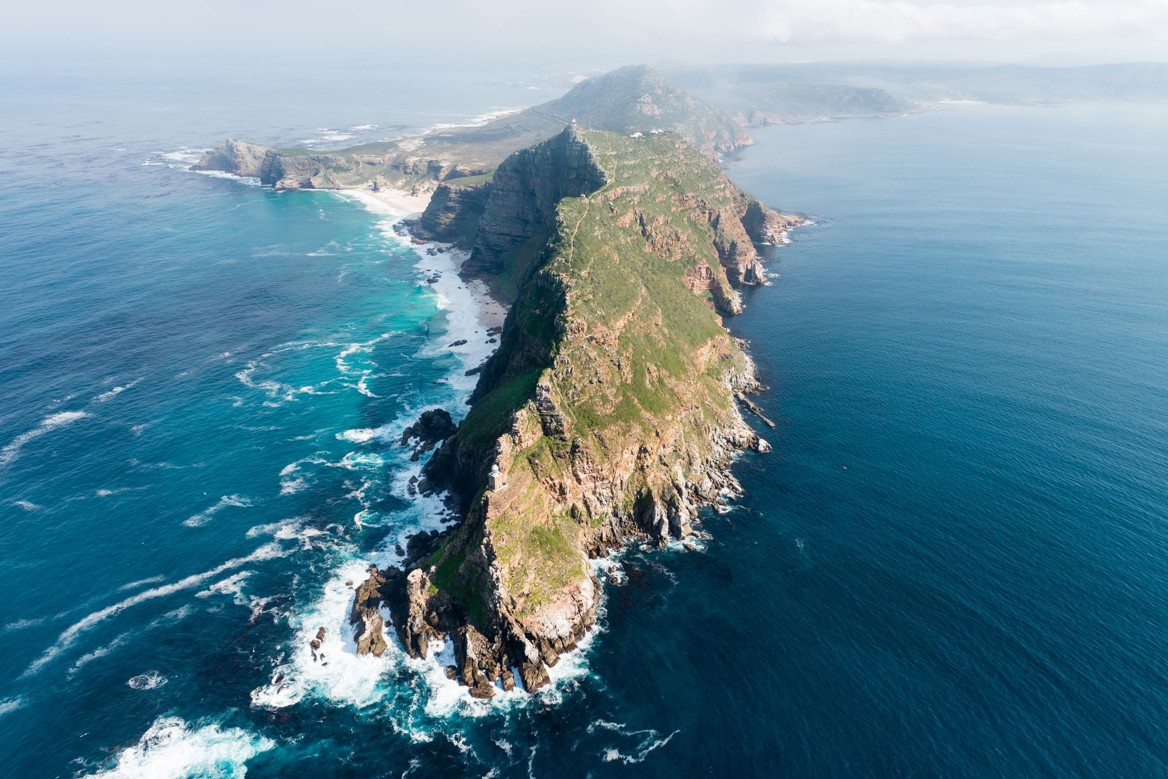 Aerial view of Cape Point Nature Reserve showcasing dramatic sea cliffs and rugged headlands covered in fynbos. The meeting of the Atlantic and Indian Oceans creates swirling waves, highlighting the raw power of nature at the edge of the African continent.