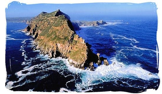 Aerial view of a rugged coastal peninsula surrounded by blue ocean waves. The rocky landmass juts out into the sea, covered with sparse vegetation. White surf breaks against the cliffs, highlighting the contrast between the land and water under a clear blue sky.