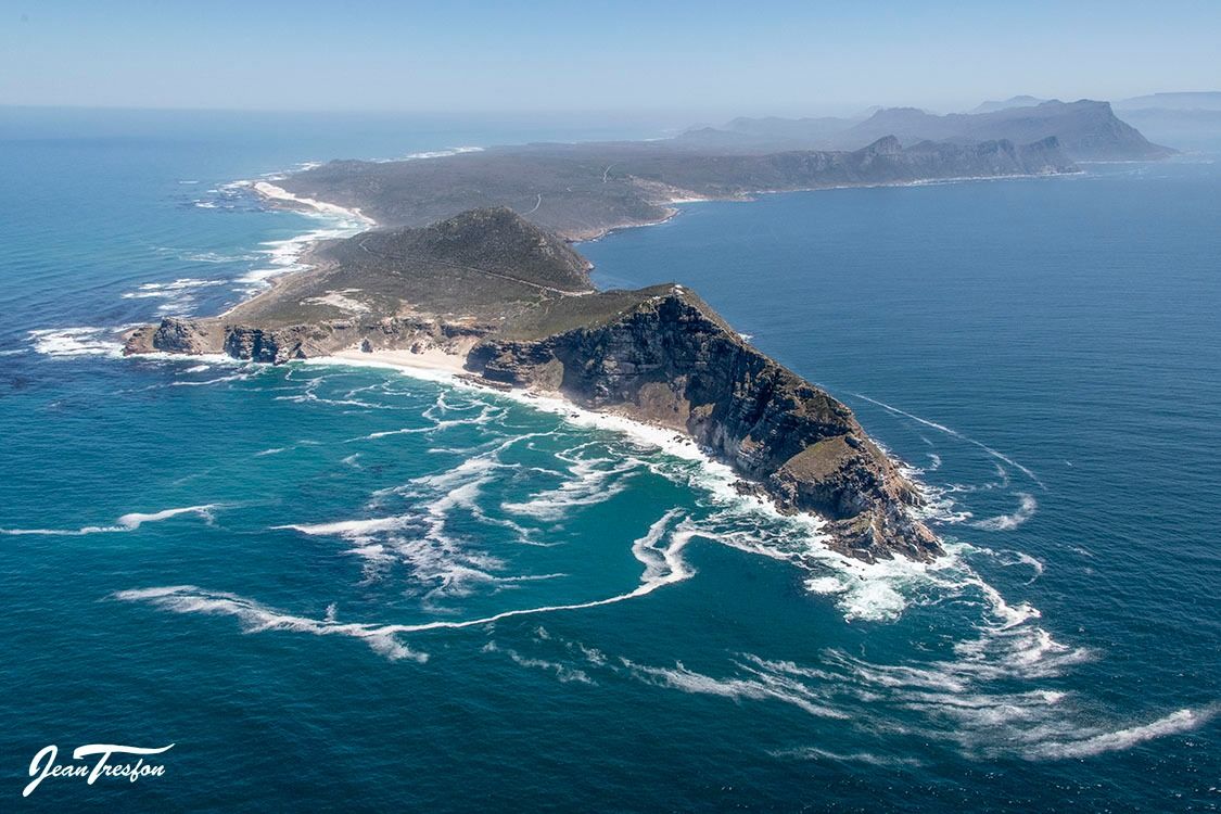 Aerial view of Cape Point Nature Reserve, featuring dramatic sea cliffs and vast ocean where the Atlantic meets the Indian Ocean. The rugged landscape is covered with fynbos, showcasing the raw natural beauty at the continent's edge. Waves crash against the rocky shores.
