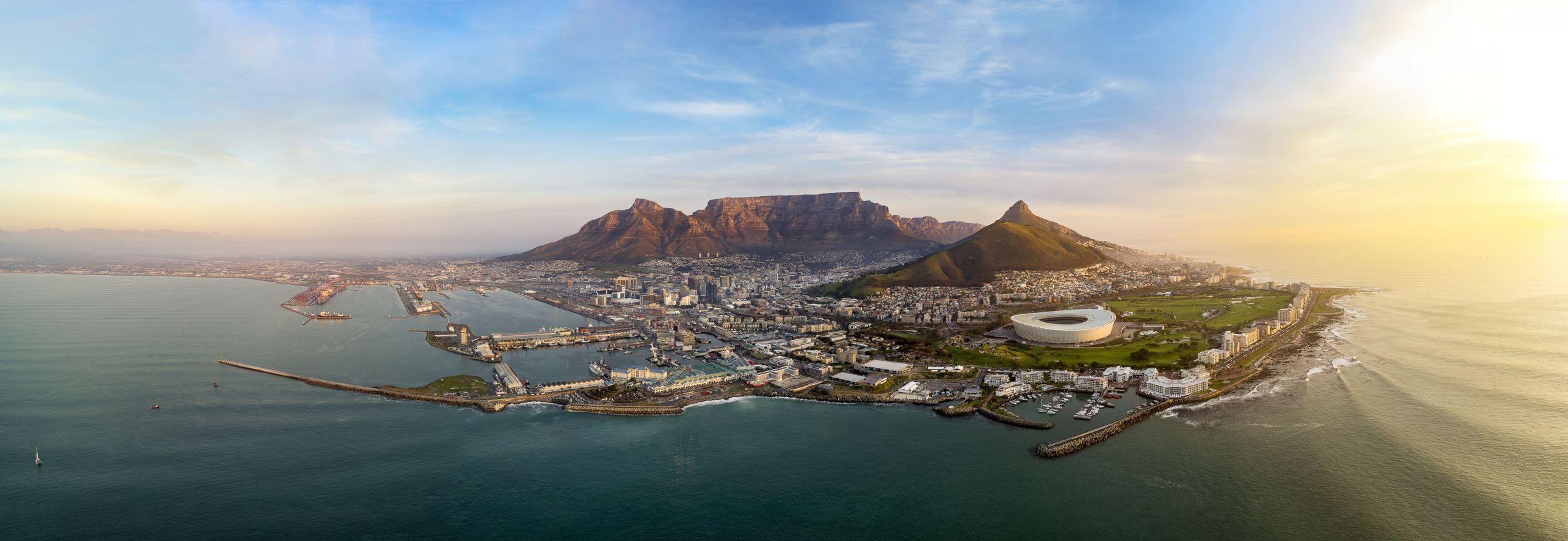 Aerial view of Cape Town, South Africa, featuring the iconic Table Mountain and Lion's Head. The cityscape includes a stadium, harbor, and coastline under a clear sky with the sun setting in the background, casting a warm glow over the scene.
