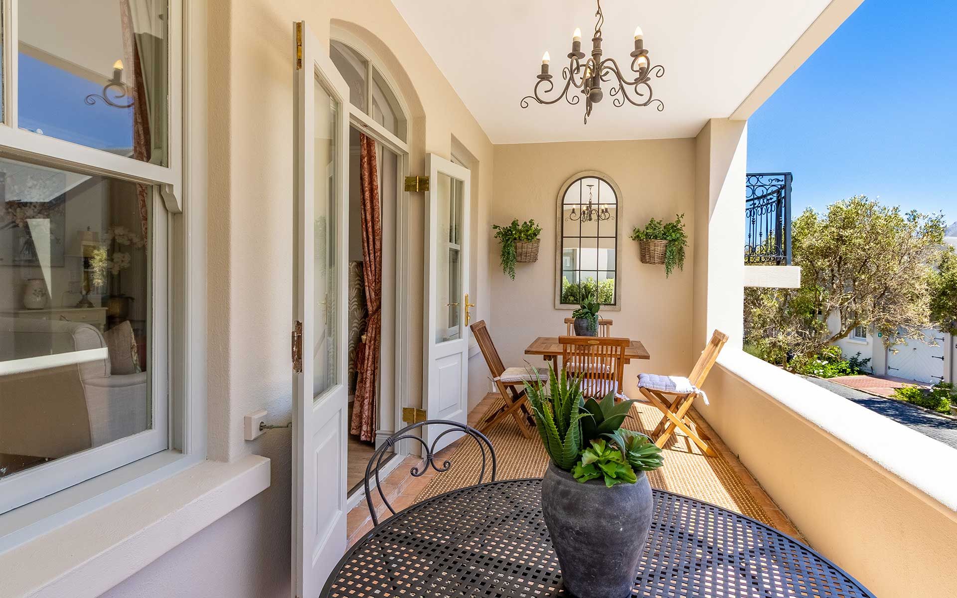 A sunlit balcony with elegant iron furniture, a wooden dining set, and lush greenery. French doors lead inside, and a chandelier adds sophistication. The view overlooks peaceful village surroundings, perfect for relaxation and enjoying nature.