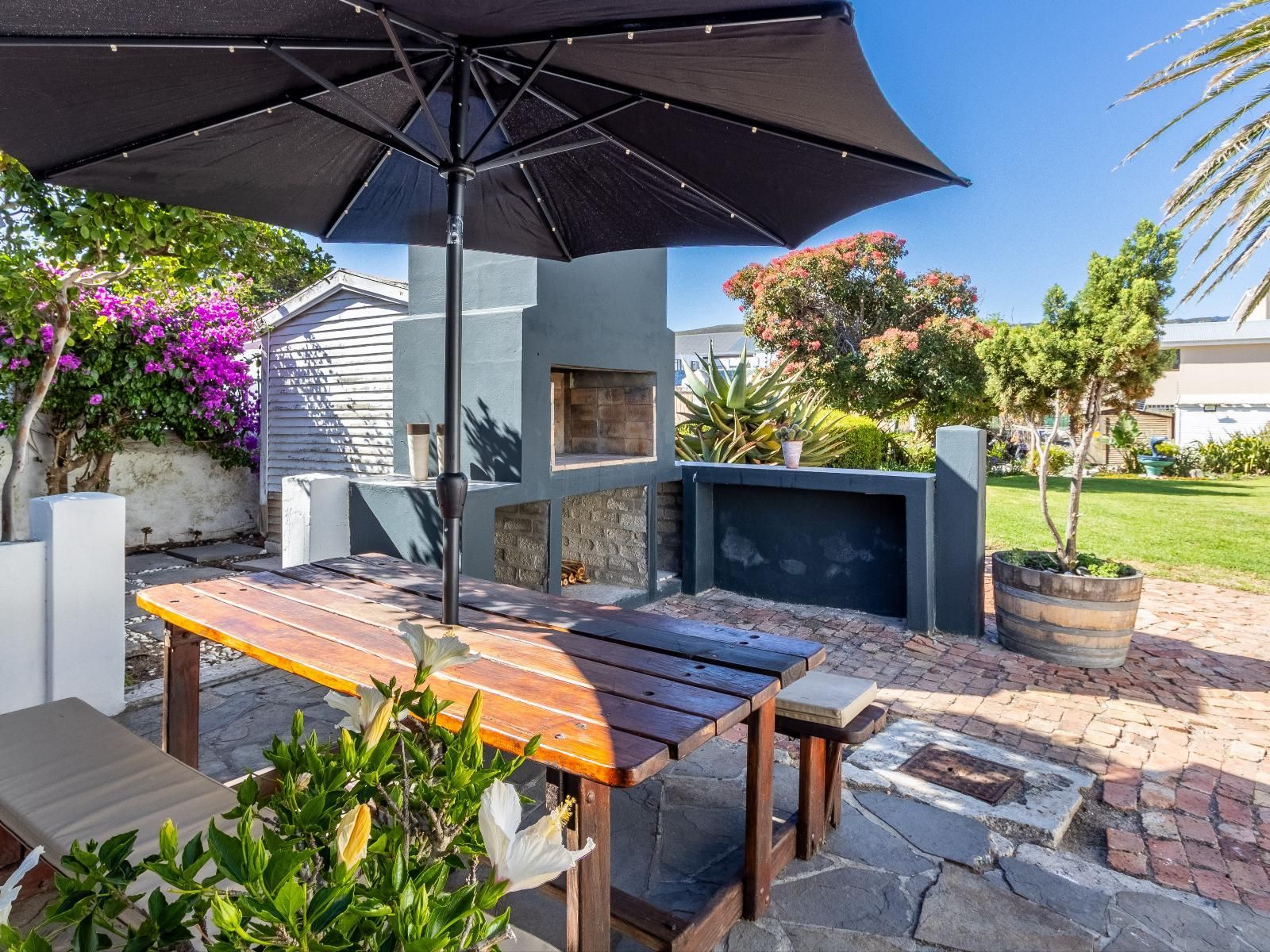 A delightful outdoor dining area featuring a wooden picnic table under a large umbrella, surrounded by lush greenery and vibrant flowers. The space includes a built-in barbecue and offers a serene garden setting perfect for alfresco meals.