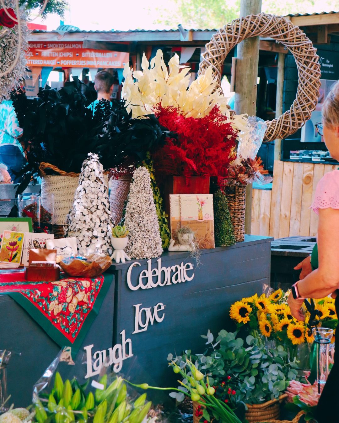A vibrant stall at Hermanus Country Market displaying a colorful array of flowers and decorative items. The words 'Celebrate', 'Love', and 'Laugh' adorn the stall, surrounded by sunflowers, festive foliage, and handmade crafts, capturing the market's lively and welcoming atmosphere.