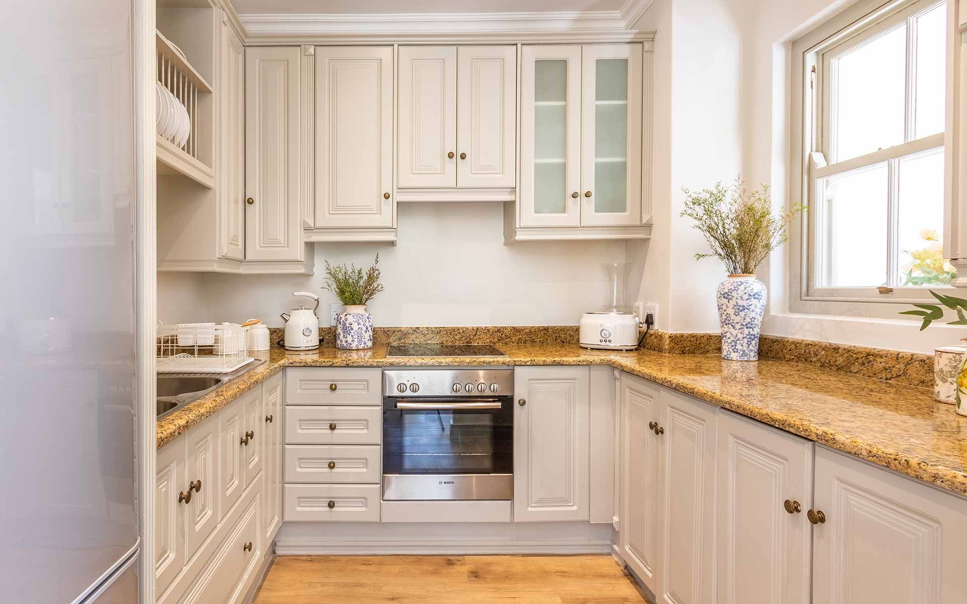 Bright kitchen with white cabinetry and granite countertops in Villa 31. Equipped with modern appliances including an oven, kettle, and toaster. Decorative vases with flowers add a homely touch. Natural light streams in through a nearby window, enhancing the inviting atmosphere.