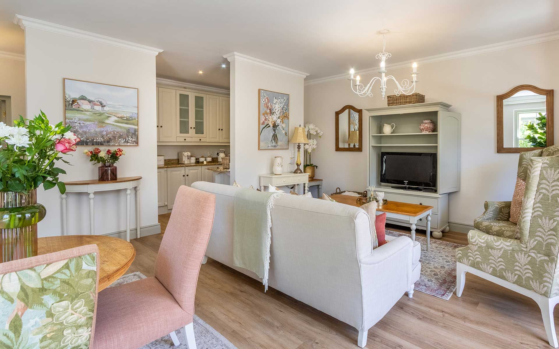 A cozy living area in Villa 27 featuring a soft beige sofa, floral armchair, and a round wooden table with pink and green upholstered chairs. The space is adorned with floral paintings, a chandelier, and a TV on a white cabinet, creating a warm and inviting atmosphere.