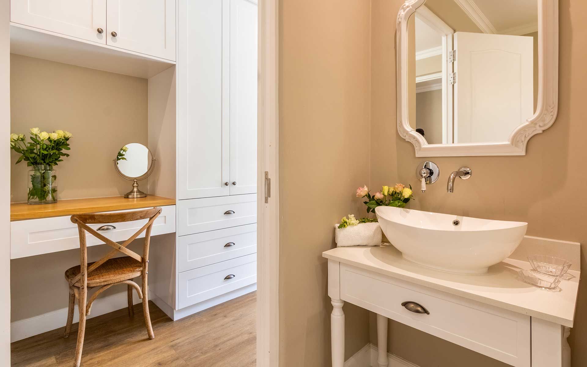 A cozy vanity area in Villa 56 featuring a wooden chair, a mirror, and a vase with fresh flowers. Adjacent is a bathroom with a white sink and elegant mirror, enhancing the villa's serene and inviting ambiance.