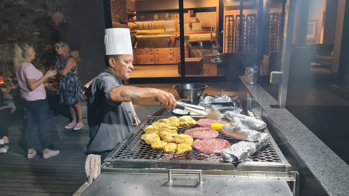 A chef wearing a white hat grills meat and corn on an outdoor barbecue. Wrapped foil packages are also on the grill. People are socializing in the background near a building with a warm interior glow.
