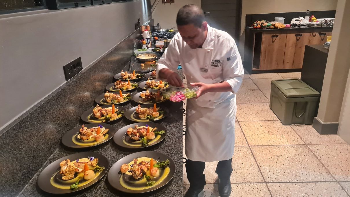 A chef in a white uniform is arranging food on several black plates lined up on a counter. Each plate is artistically arranged with vegetables and meat. The kitchen setting features a granite countertop and various kitchen tools in the background.