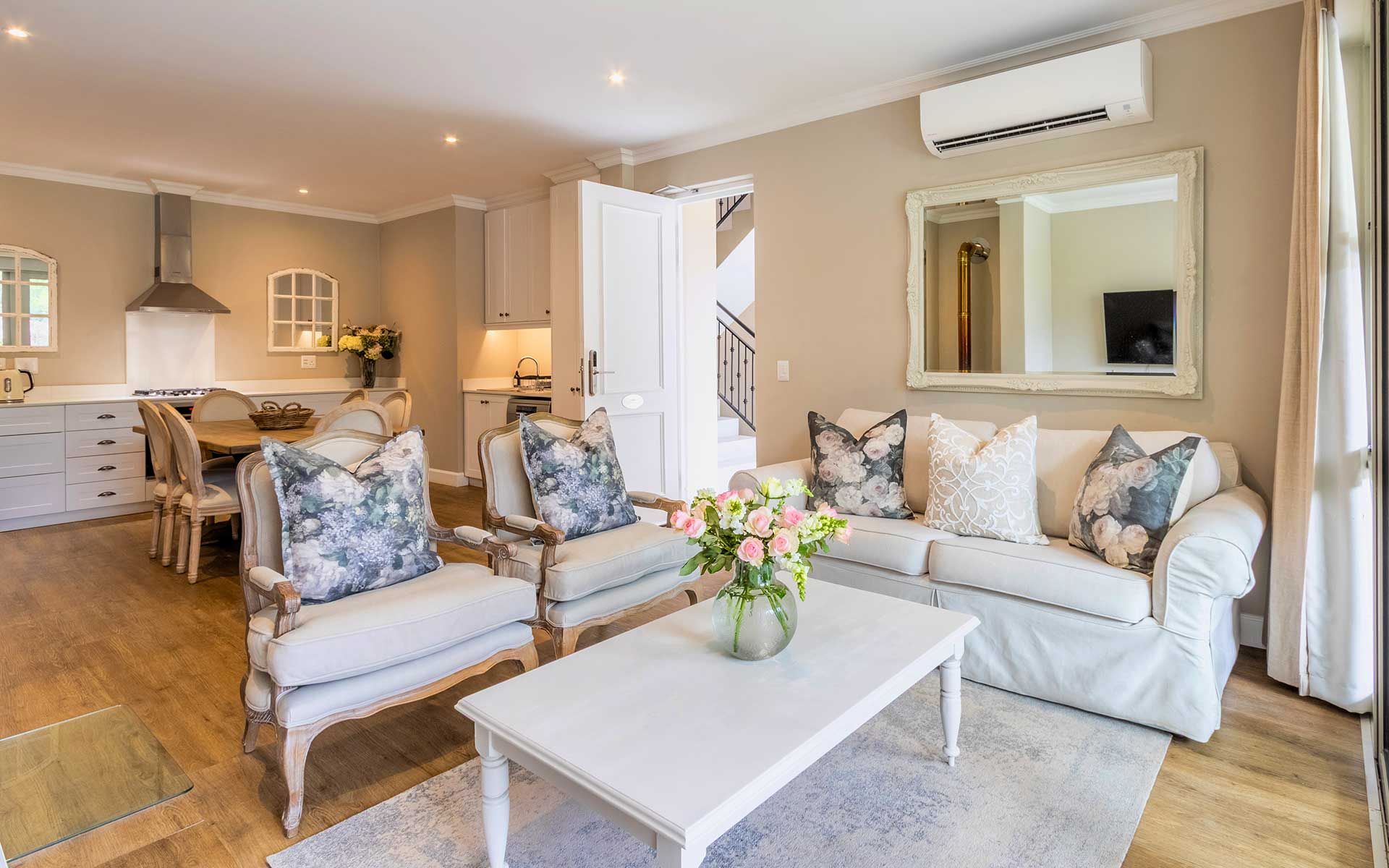 A stylish villa living area with elegant furniture, including a white sofa and armchairs with floral cushions. A white coffee table with fresh flowers complements the space. The open-plan design connects to a modern kitchen, enhancing the villa's inviting and cozy atmosphere.