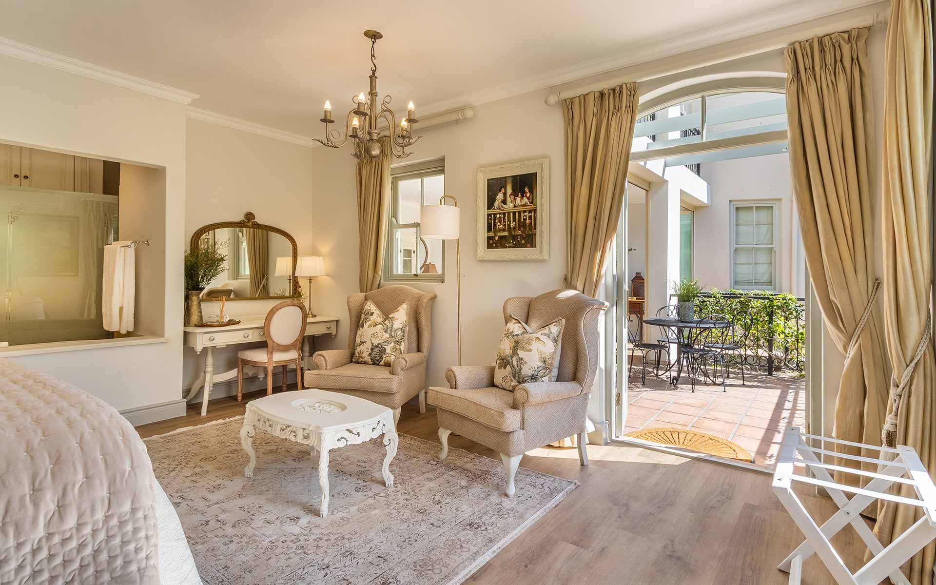 A luxurious villa interior with elegant armchairs, a vintage vanity, and a chandelier. Natural light filters through draped curtains, leading to a quaint patio with wrought-iron furniture. The setting exudes warmth and sophistication, ideal for a serene retreat.