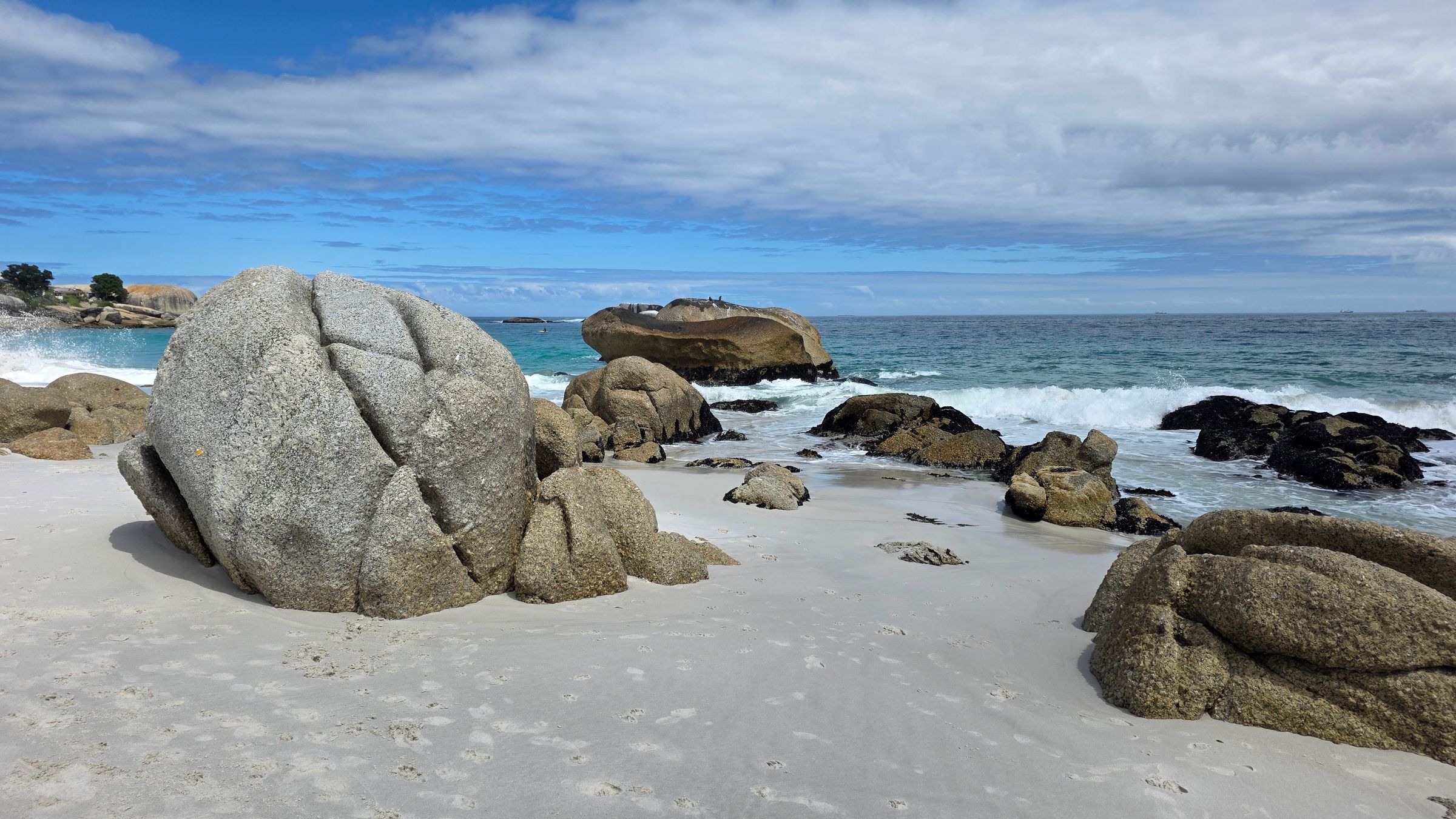 A scenic view of Cape Town's Clifton Beach featuring large boulders on white sand with turquoise Atlantic waves crashing gently. The image captures the pristine natural beauty and dramatic coastal scenery under a partly cloudy sky.