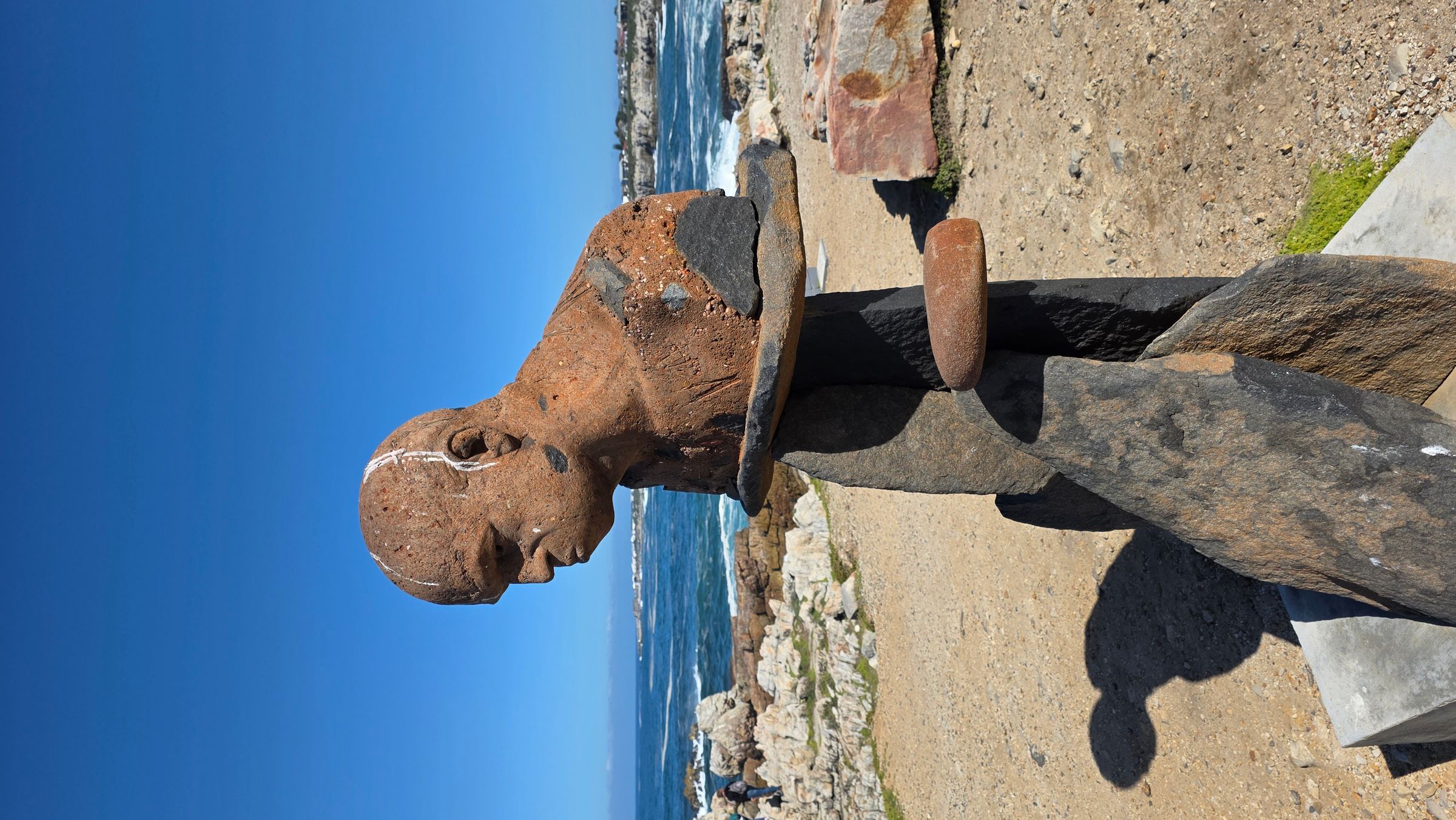 A stone sculpture of a human figure, blending with the rocky coastal backdrop in Hermanus. The artwork features a textured, earthy finish and is positioned against a clear blue sky, capturing the essence of the town's artistic and natural beauty.