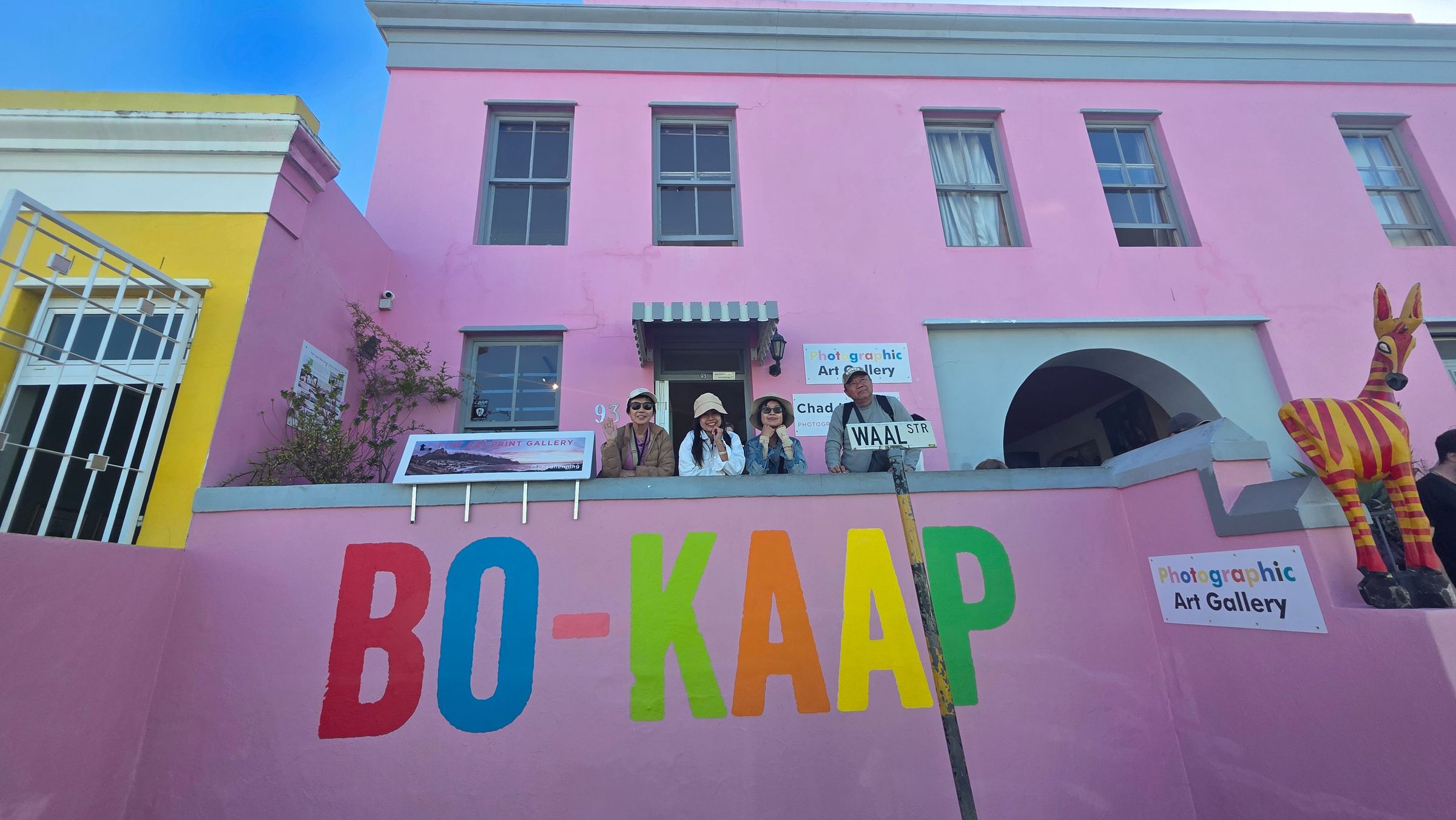 A vibrant pink building in Bo-Kaap, Cape Town, featuring bold 'BO-KAAP' lettering in multiple colors. People stand on the balcony above a photographic art gallery sign. A yellow and pink striped animal sculpture adds to the lively scene, capturing the area's cultural charm.