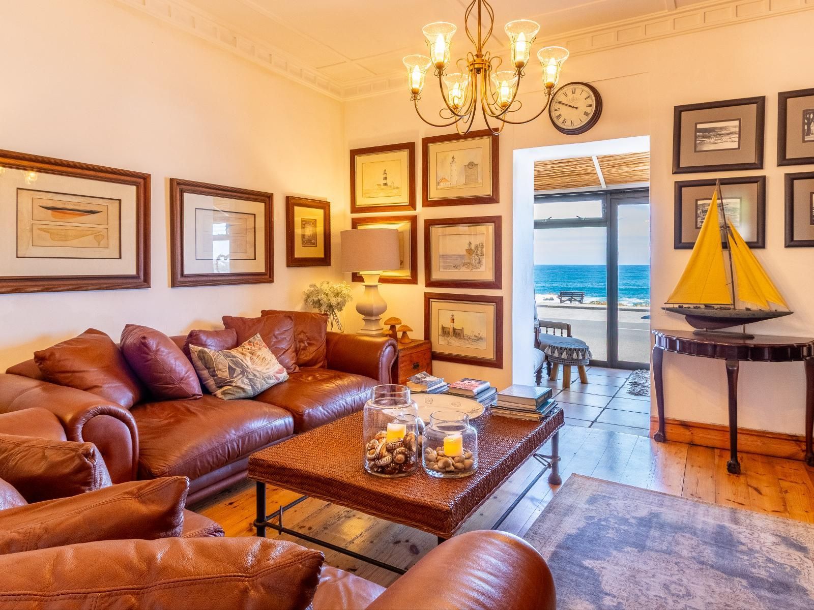 The lounge features plush leather sofas, nautical-themed decor, and a coffee table. A wide doorway reveals a sunlit patio with a stunning ocean view, perfect for relaxing. Elegant lighting and framed art add to the room's charm and warmth.