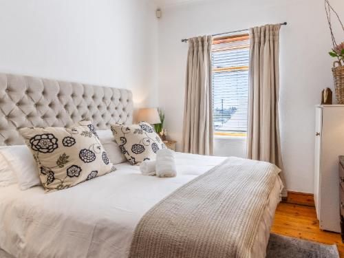A bedroom with a tufted headboard, patterned pillows, and beige curtains. Soft lighting enhances the natural light from the window. The room has a wooden floor and a warm, inviting atmosphere with simple, elegant decor.
