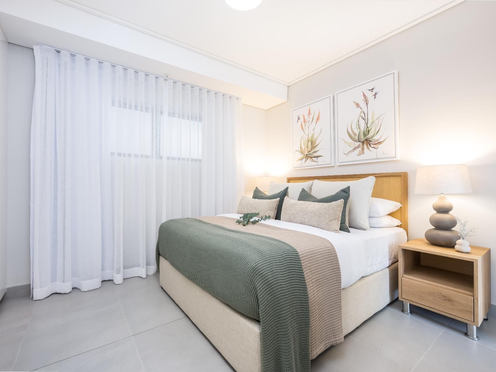 A serene bedroom with a double bed adorned with green and beige bedding, flanked by a modern wooden nightstand and lamp. The room features two botanical wall prints and sheer curtains, creating a calm and inviting atmosphere.