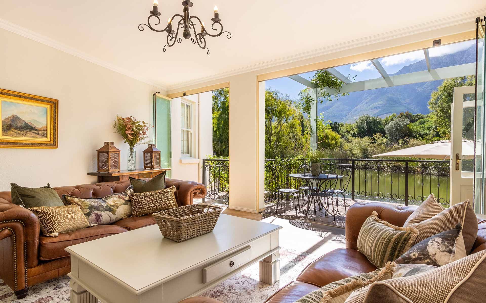 A cozy villa living area with leather sofas and patterned cushions. A table centerpiece and chandelier add charm. Open doors lead to a balcony with wrought-iron furniture, overlooking a serene lake and lush mountains in the distance.