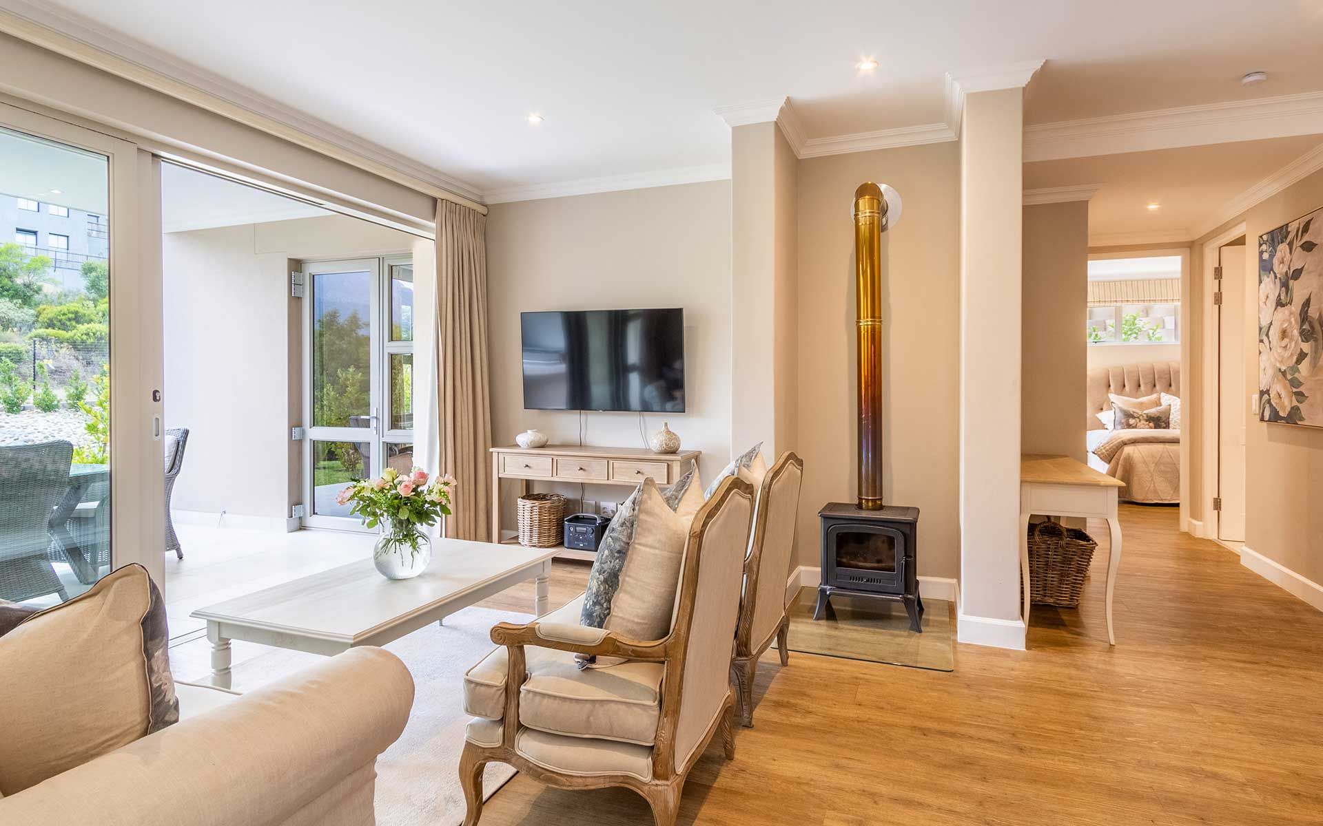 A bright and cozy living room in the villa, featuring elegant furnishings and a wall-mounted TV. Large glass doors open to a covered patio with garden views. A wood-burning stove adds warmth beside a hallway leading to a sunlit bedroom with plush bedding.