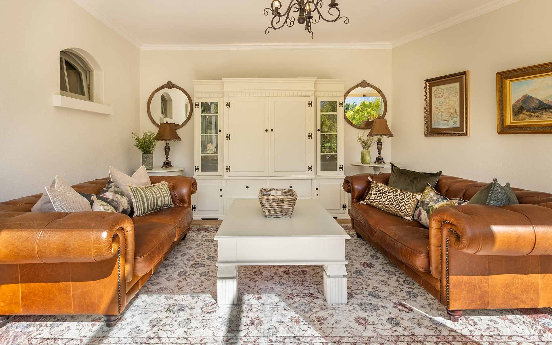 A warm lounge with brown leather sofas, decorative cushions, and a white coffee table. The room features a white cabinet, mirrors, and soft lighting from elegant lamps. The cozy ambiance is enhanced by natural light and a patterned rug.