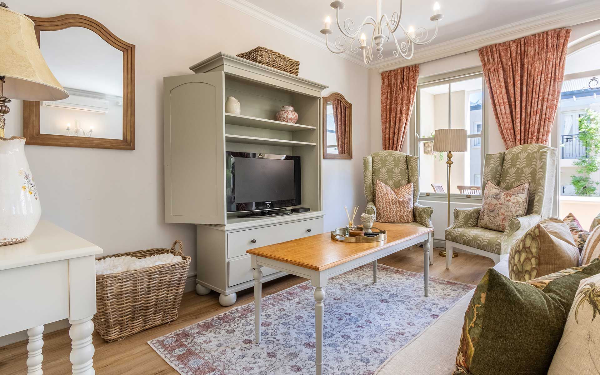 A charming living room with elegant decor, featuring two patterned armchairs, a wooden coffee table, and a TV cabinet. Soft lighting from a chandelier and natural light from large windows create a warm ambiance. Decorative pillows and a woven basket add to the cozy atmosphere.
