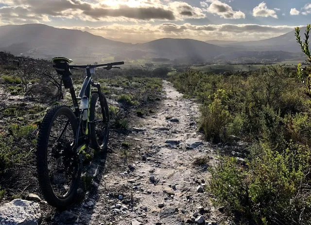A mountain bike rests on a rocky trail surrounded by greenery, with a scenic view of distant mountains under a cloudy sky. The sun casts rays through the clouds, illuminating the landscape in the background.