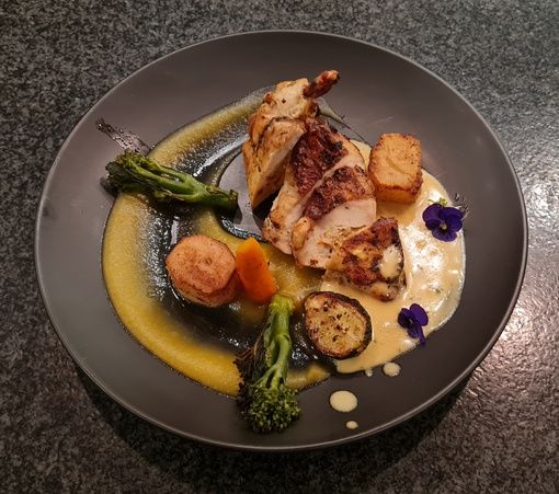 A beautifully plated dish featuring sliced grilled chicken breast, two pieces of fried polenta, and an assortment of vegetables including broccolini, carrot, and zucchini. The dish is garnished with edible flowers and drizzled with a creamy yellow sauce, served on a dark plate.