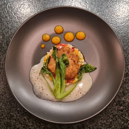 A gourmet dish featuring a crispy round breaded item atop a bed of colorful vegetables, garnished with green leaves. Accompanied by a creamy white sauce and circular dollops of orange puree, elegantly arranged on a dark plate with a textured background.