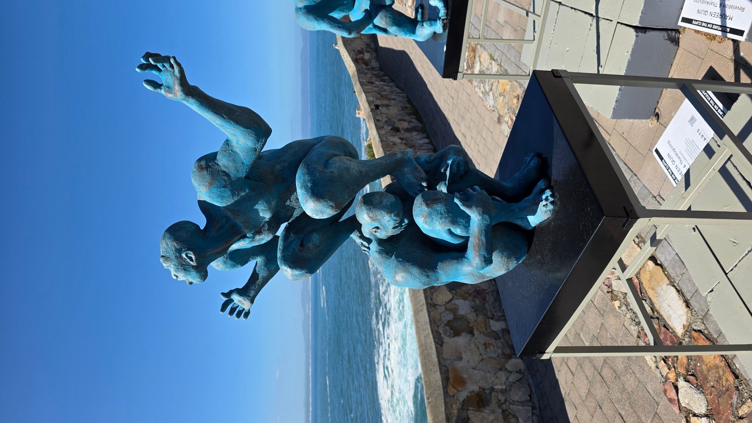 A striking blue sculpture depicting intertwined figures, set on a coastal promenade in Hermanus. The artwork captures a sense of movement against a backdrop of ocean and clear sky, highlighting the town's vibrant arts scene and outdoor exhibitions.