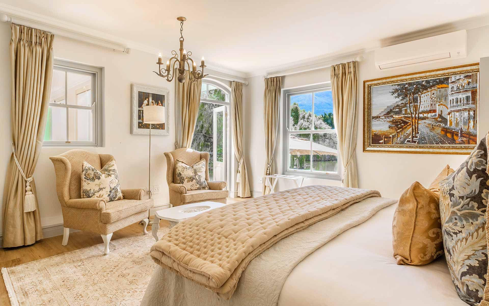 Luxurious bedroom with a king-size bed adorned with plush pillows and a quilted throw. Two armchairs sit by large windows with scenic views of a garden and mountains. Soft, neutral tones and elegant decor create a serene atmosphere, highlighted by a classic chandelier and framed artwork.
