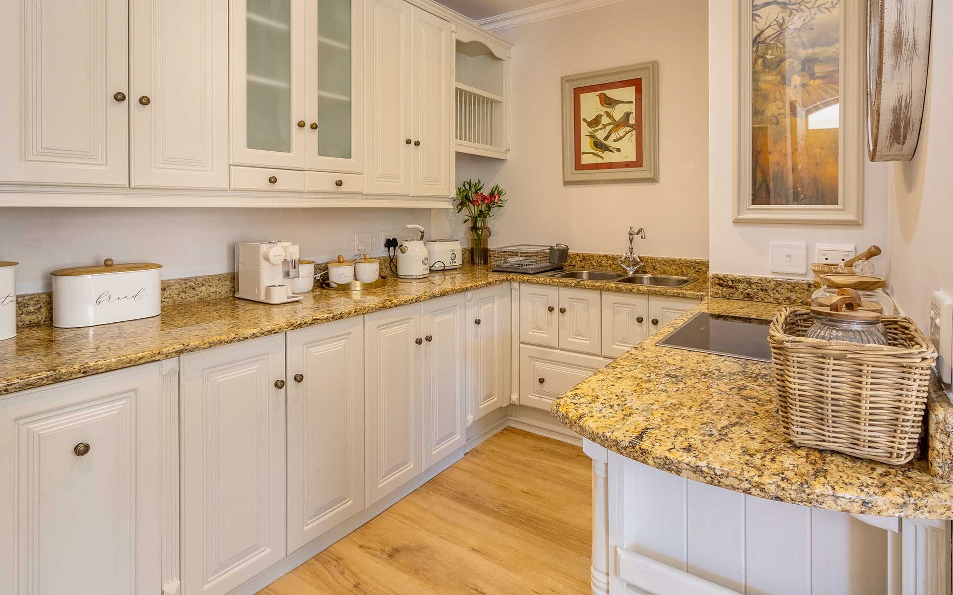 A charming kitchen with granite countertops, white cabinetry, and modern appliances. Accents include a coffee maker, toaster, and decorative jars. A framed bird illustration adds warmth, complementing the wooden floor for a cozy, stylish culinary space.