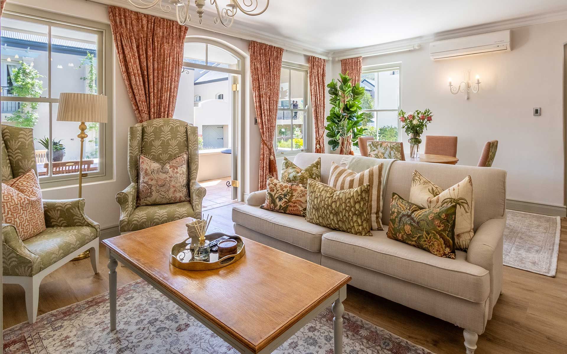A cozy living room with plush seating, patterned cushions, a wooden coffee table, and floral decor. Sunlight streams through large windows with coral curtains, highlighting the stylish ambiance, ideal for relaxation.