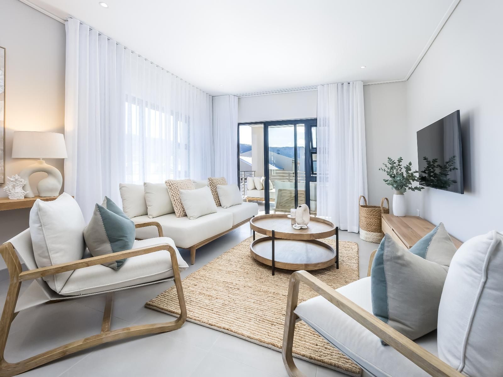 A stylish living room featuring plush white sofas, wooden accents, and a round coffee table. Soft natural light filters through sheer curtains. A flat-screen TV is mounted on the wall. The space opens to a balcony with outdoor seating, offering a view of distant hills.