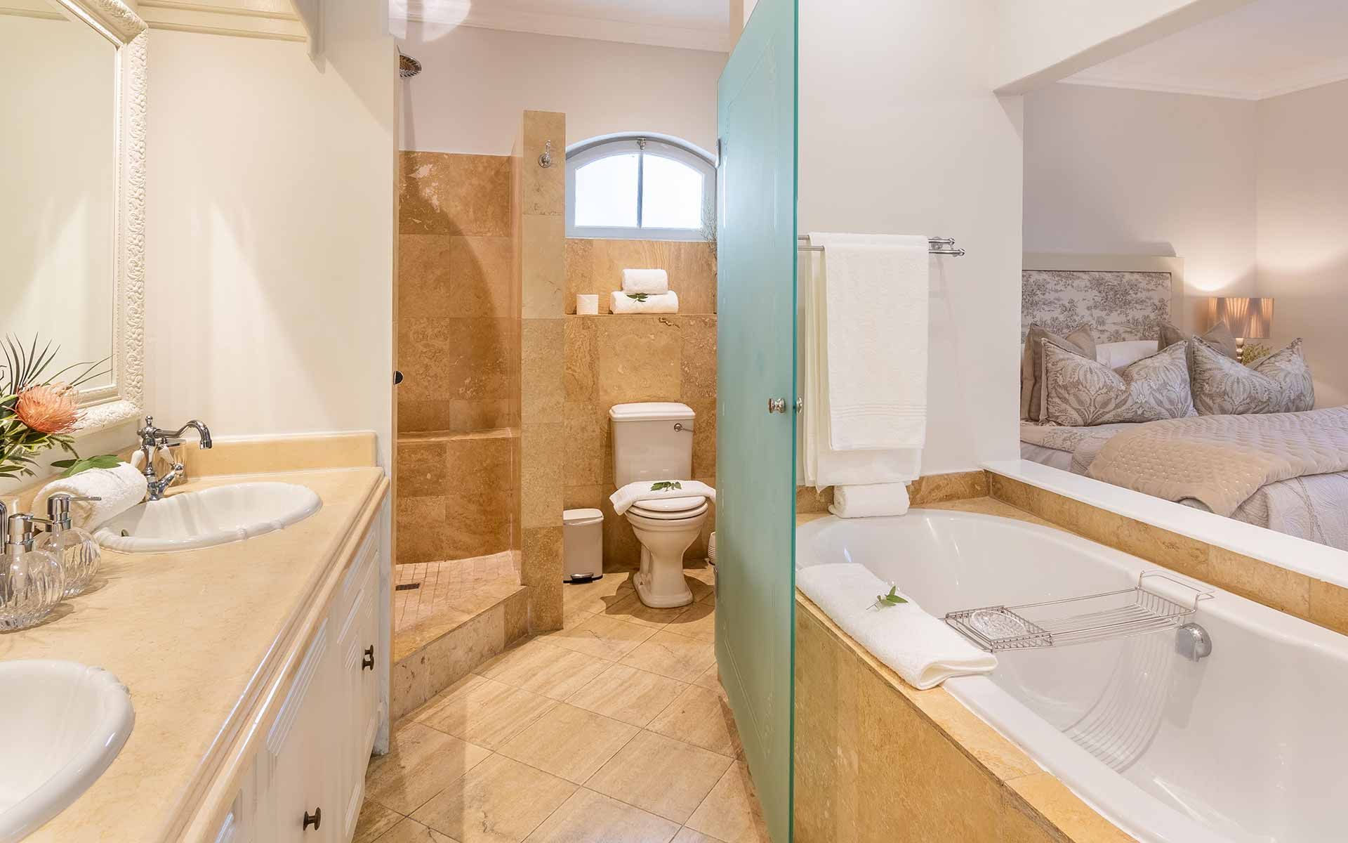 A luxurious bathroom featuring a double vanity with a large mirror, a spacious shower, and a bathtub adorned with plush towels. The light tones and natural materials create a serene and inviting atmosphere, with a glimpse into a tastefully decorated bedroom adding to the villa's charm.