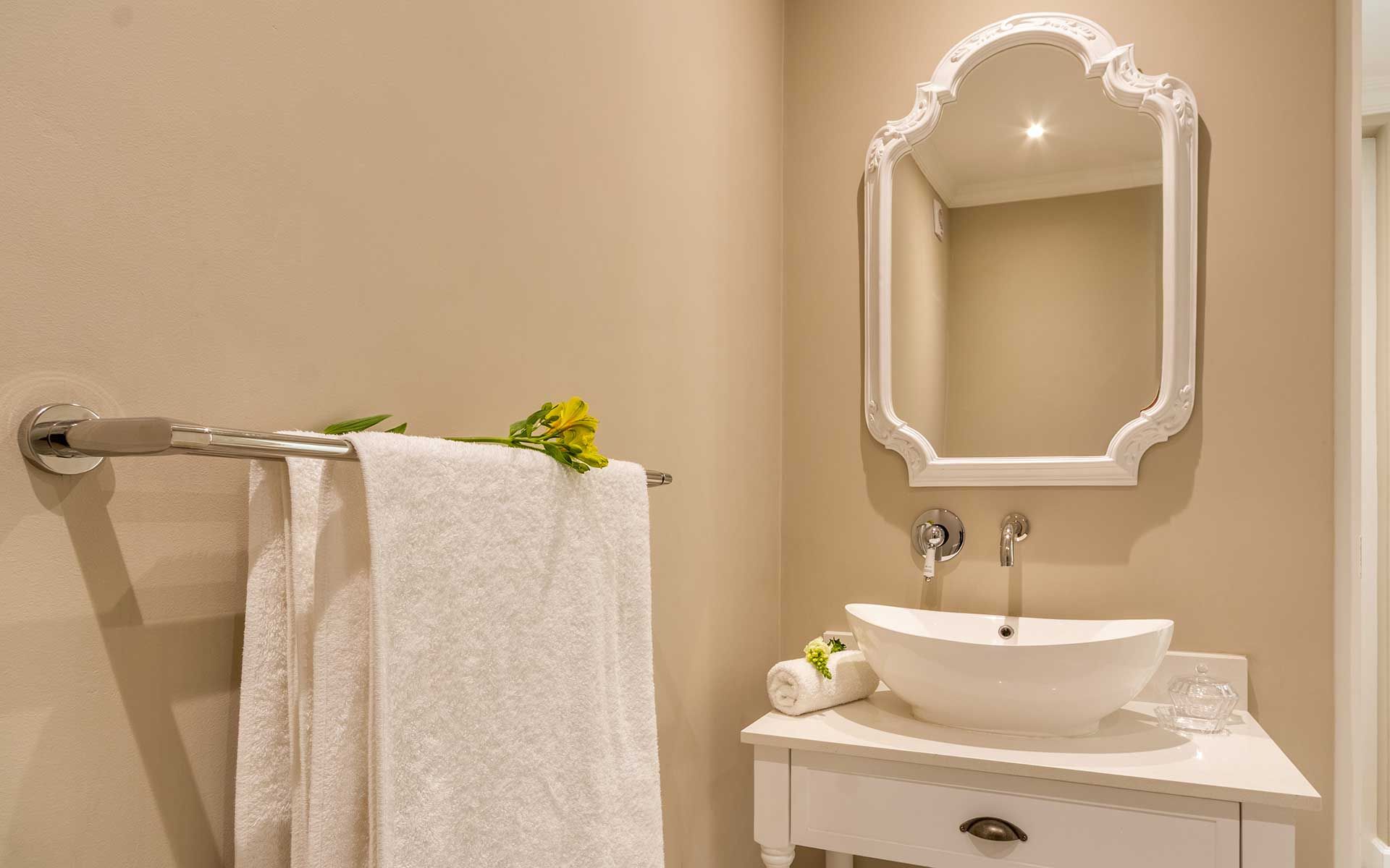 A chic bathroom in Villa 56 featuring a white, ornate-framed mirror above a modern vessel sink atop a sleek white vanity. A towel with a yellow flower rests on a chrome towel bar, complementing the serene, neutral-toned walls and soft lighting.