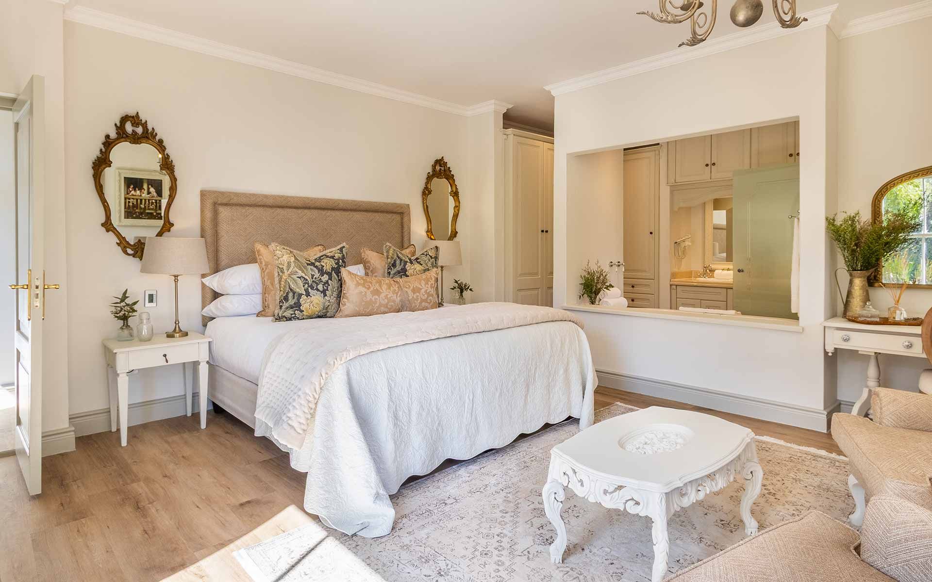 A spacious, elegantly decorated bedroom with a king-size bed adorned with patterned pillows and a plush headboard. The room features ornate mirrors, a chandelier, and a cozy seating area, offering a luxurious and tranquil retreat with soft, natural lighting enhancing the serene ambiance.