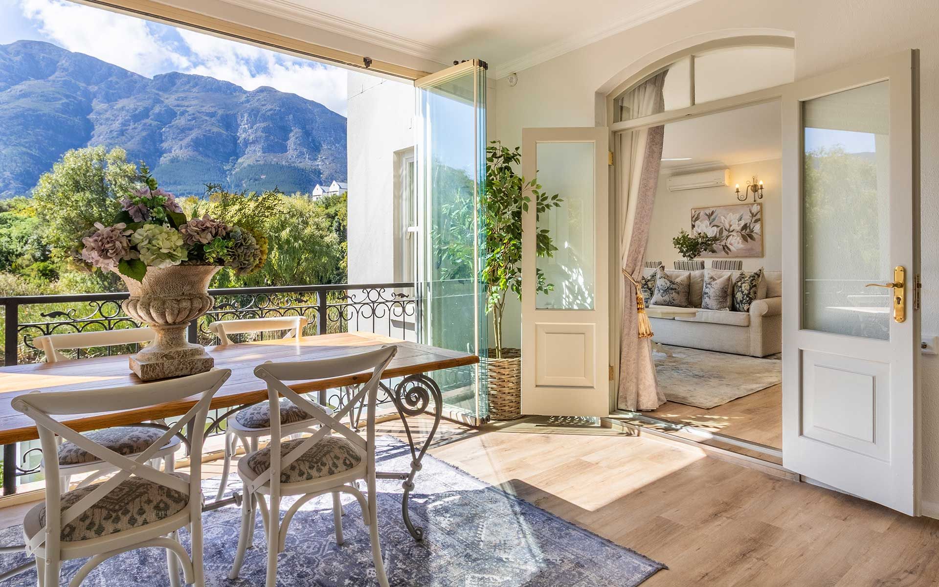 A bright dining area with a wooden table and cushioned chairs overlooks scenic mountain views. Large glass doors open to a cozy living room with a sofa and decorative plants, creating a seamless blend of indoor and outdoor spaces in Villa 31, Franschhoek.